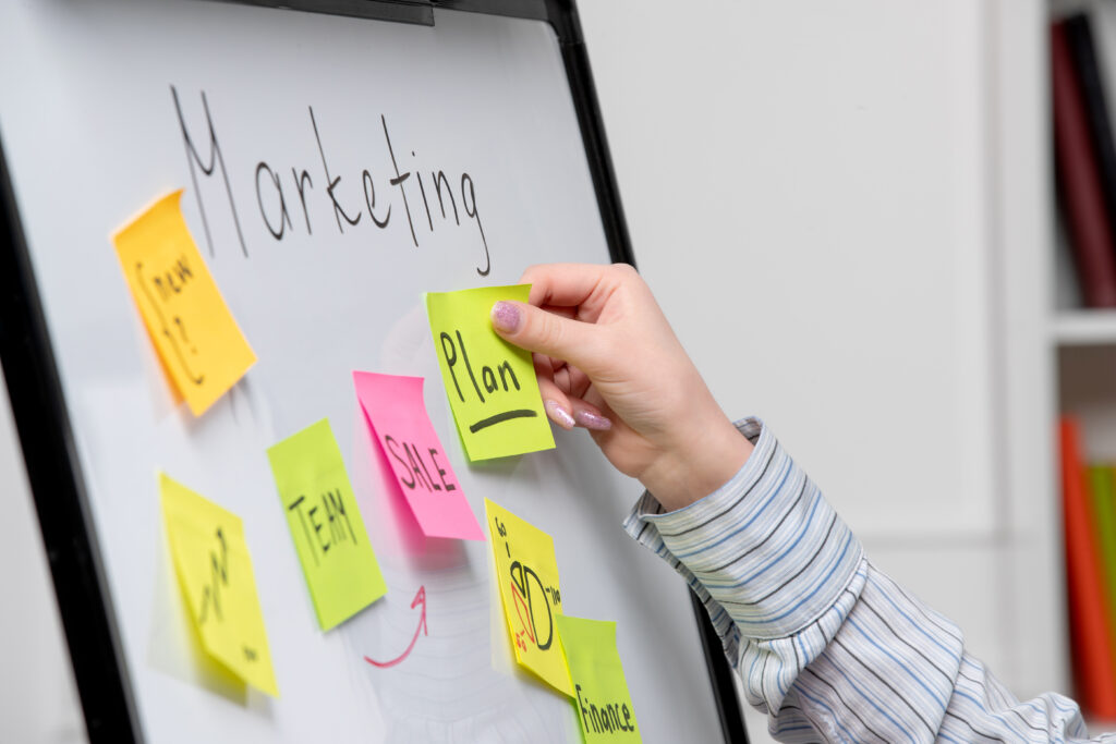 Pessoa organizando ideias em um quadro branco com post-its coloridos para desenvolver um plano de marketing estratégico.