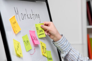 Pessoa organizando ideias em um quadro branco com post-its coloridos para desenvolver um plano de marketing estratégico.